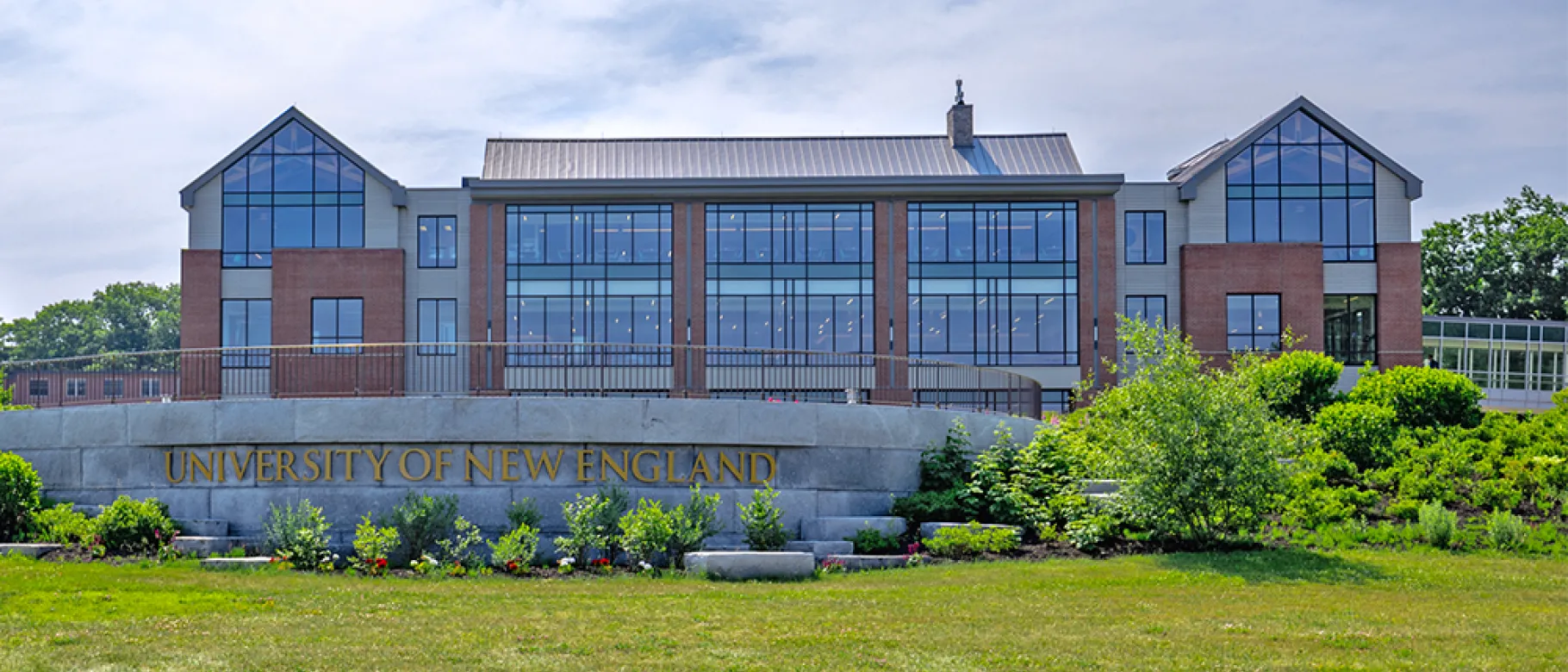 A brick building with numerous windows sits behind a stone sign that reads, "University of New England"