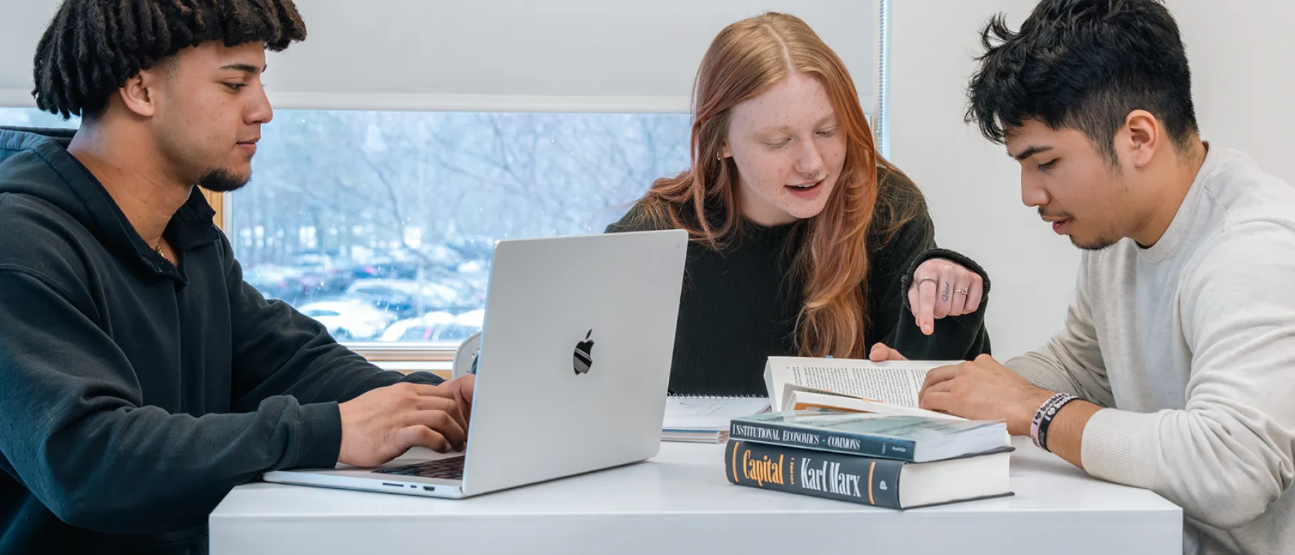 A student types on their laptop while two students review a book while sitting together at a high-top desk in front of a large window