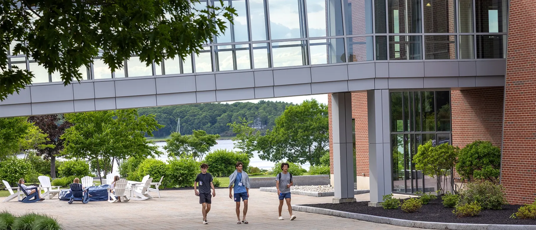 Three students wearing lanyards walk across a sunny brick courtyard beneath a glass pedestrian bridge connecting campus buildings overlooking the water.