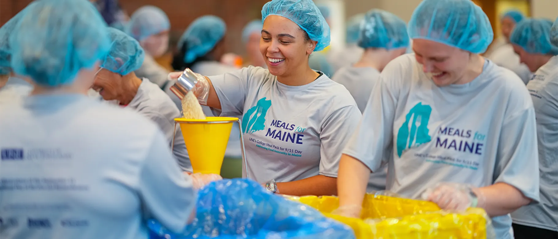 Volunteers in hairnets smile while packing bags of food at U N E's 2025 Meals for Maine event addressing food insecurity.