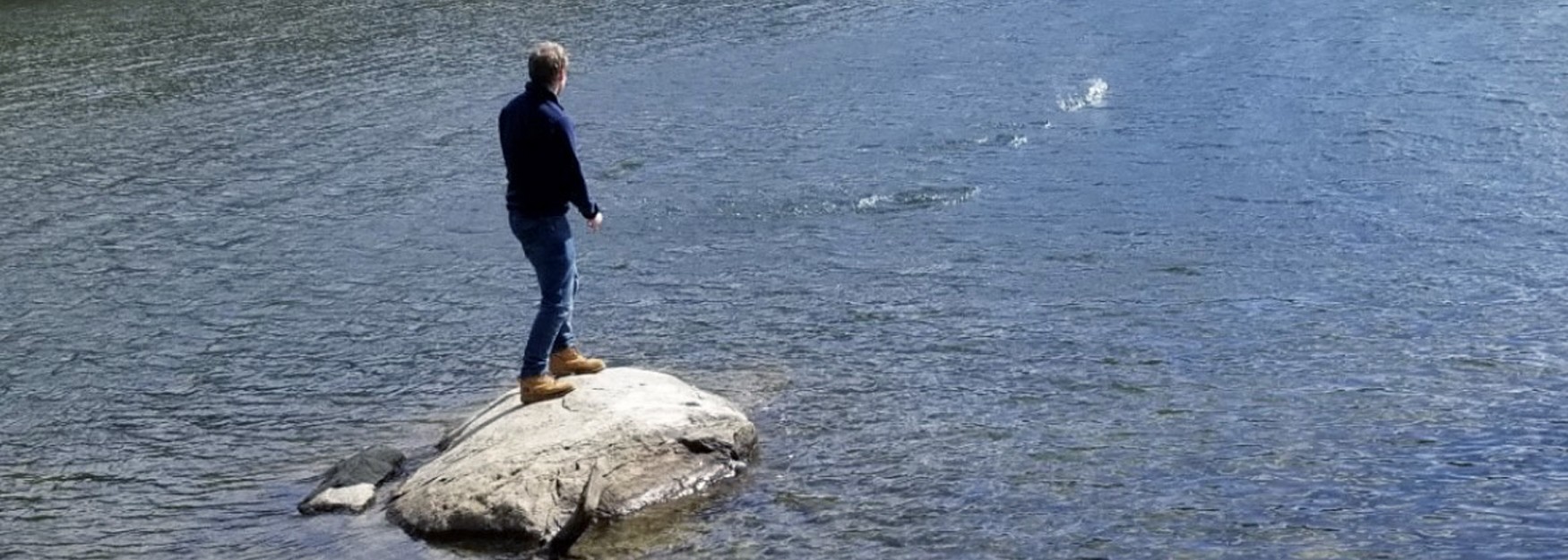 Luke Colomey stands on a rock in a body of water, his back facing the camera