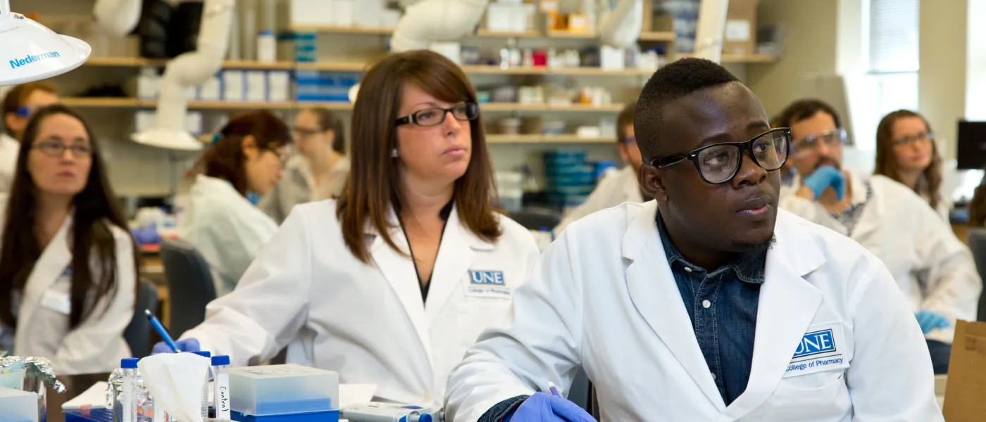 U N E College of Pharmacy students wearing lab coats take notes