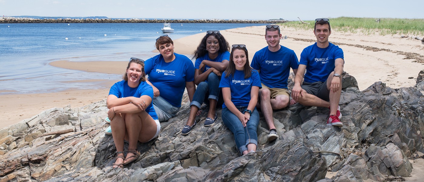A group of U N E student tour guides wear matching blue shirts while sitting on a rocky beach