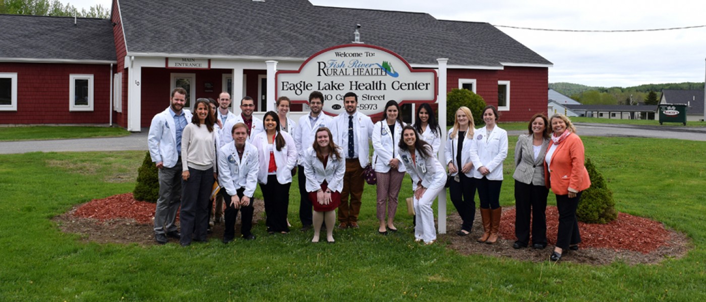 A group of U N E Health professions students pose in white coats in front of the sign for Fish River Rural Health Center