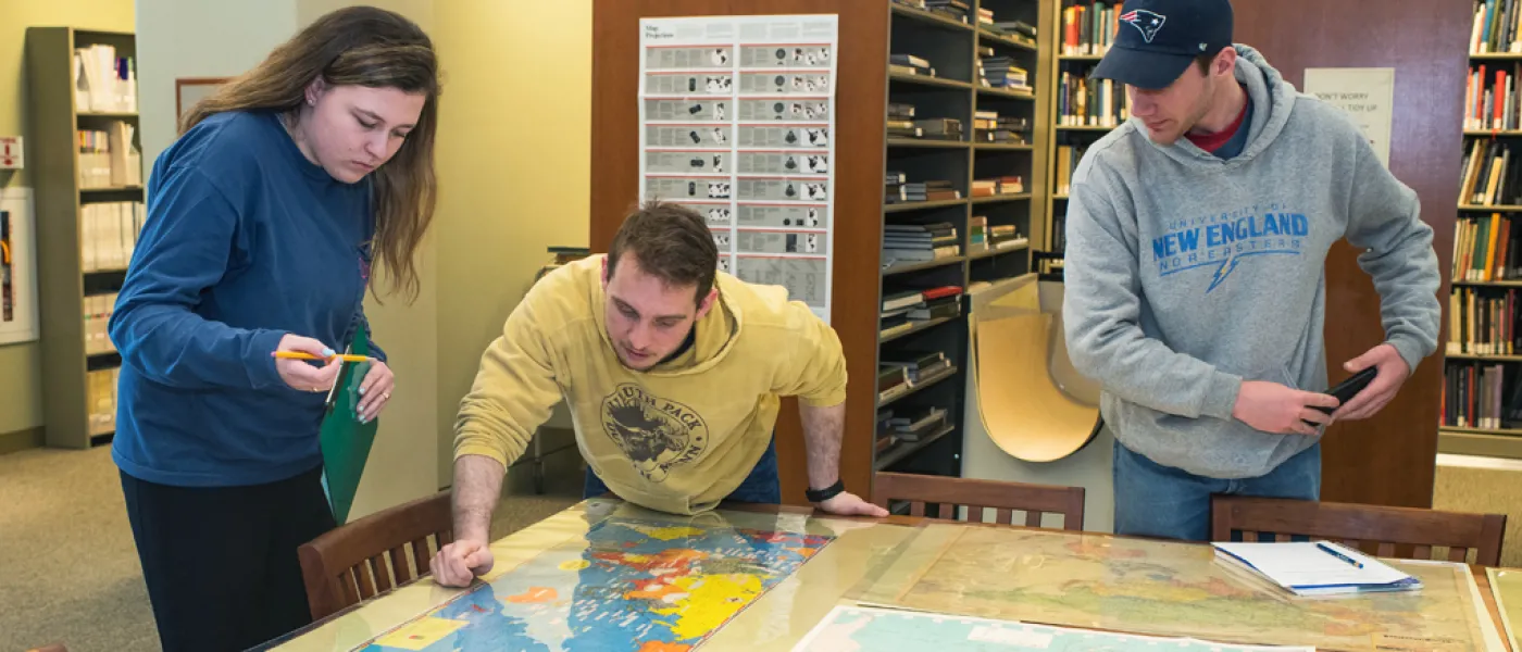 Three U N E Liberal Arts students study a large map in the library