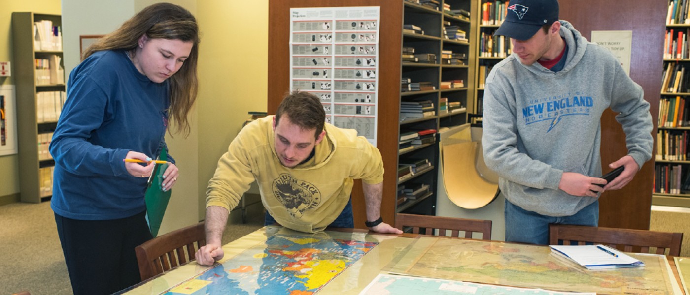 Three U N E Liberal Arts students study a large map in the library