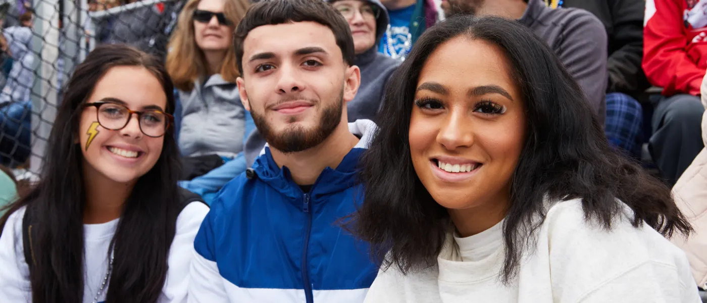 Three students at a U N E football game smile at the camera