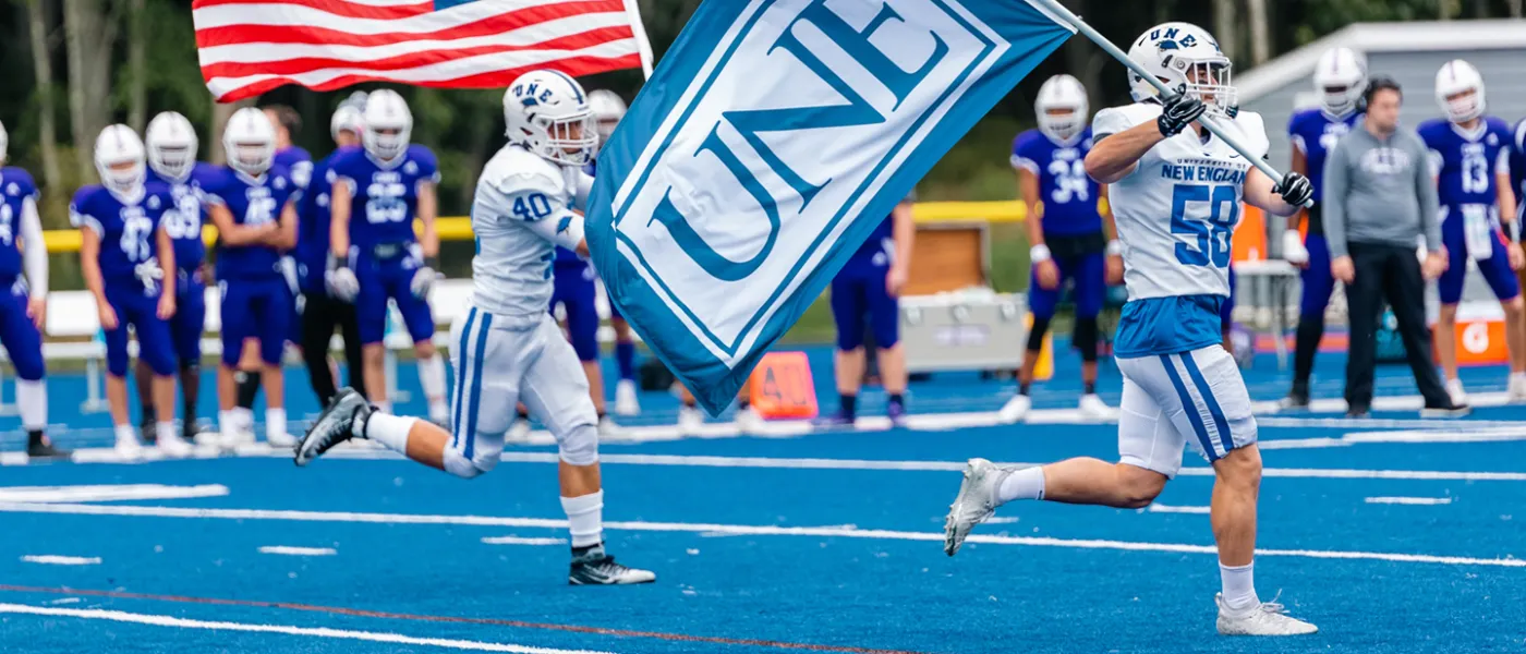 Members of the U N E football team running onto the field with an American flag and a U N E flag