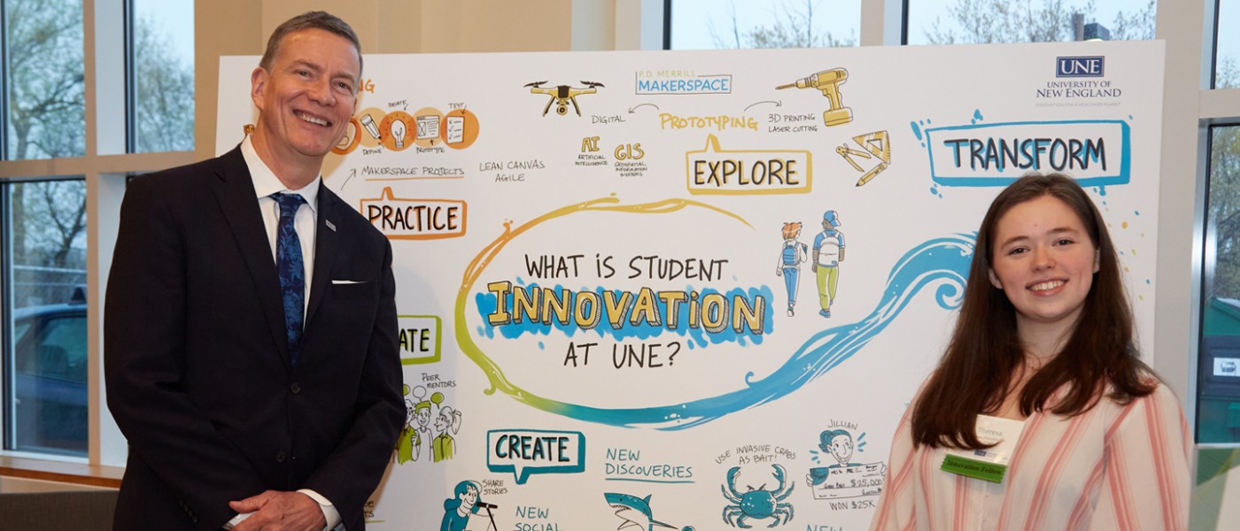 President Herbert stands with a U N E student in front of the Student Innovation poster
