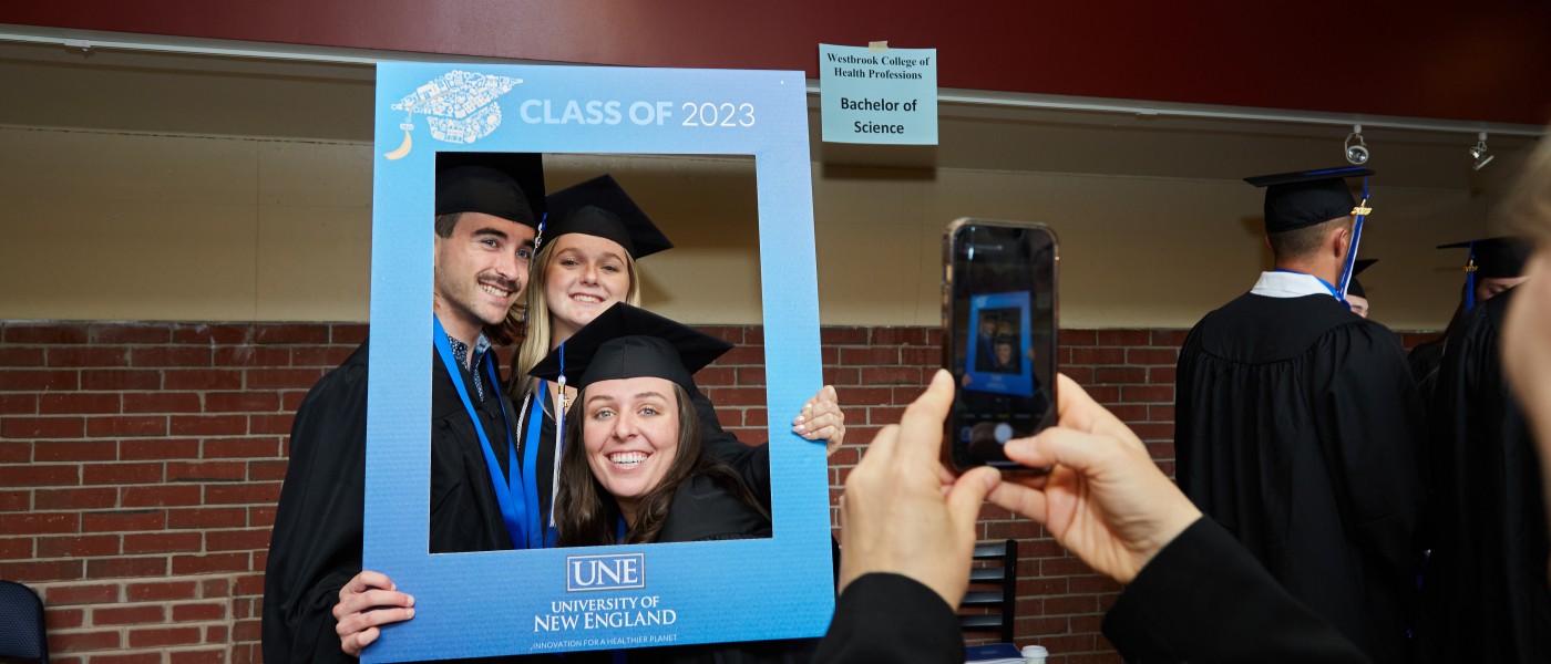 Students dressed in cap and gown pose for a photo with an oversized frame