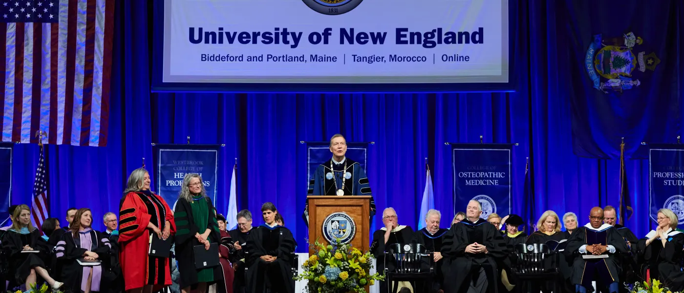 U N E President james herbert stands at the podium with faculty seated behind him at 2023 commencement
