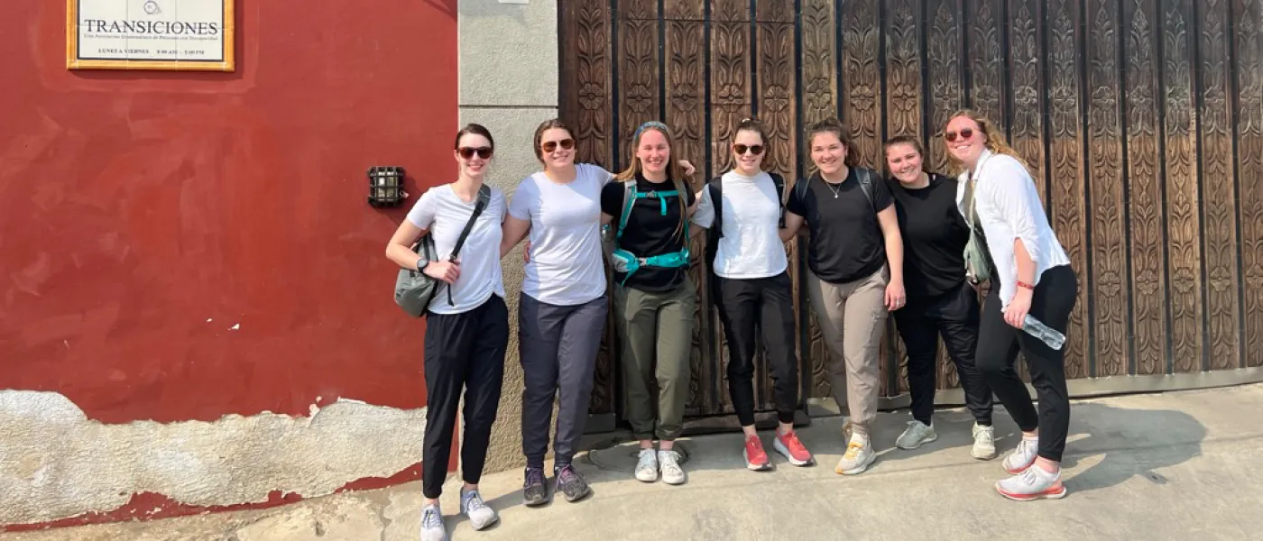 Seven friends stand in a row in front of a red building with a decorative large wooden door