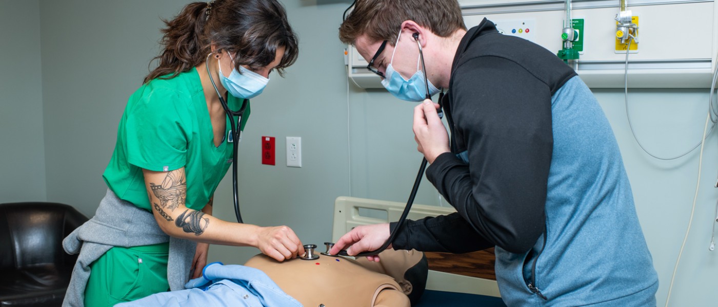 Two physician assistant students use stethoscopes to listen to the heartbeat of a patient simulator