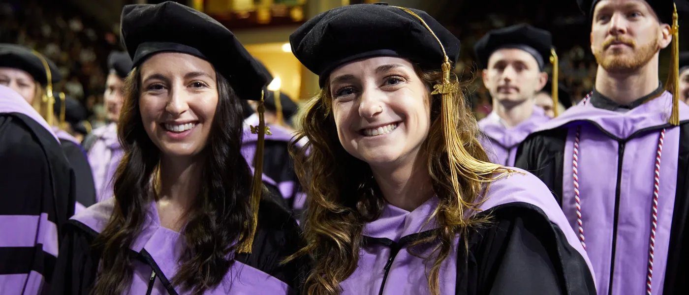 A group of graduate students in purple robes smile at the camera at graduation