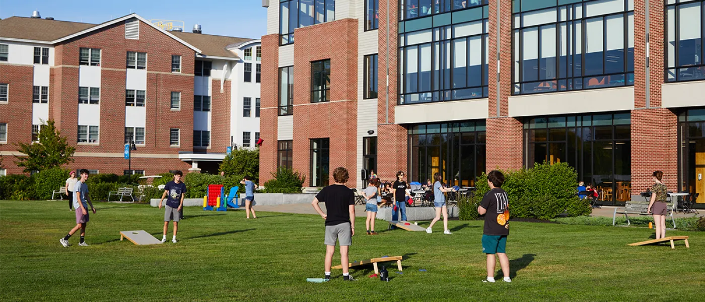 A group of students playing corn hole on green grass in front of two large brick buildings with numerous windows