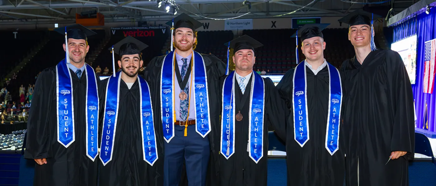 A group of graduating students pose in regalia with blue stoles that read "student athlete"
