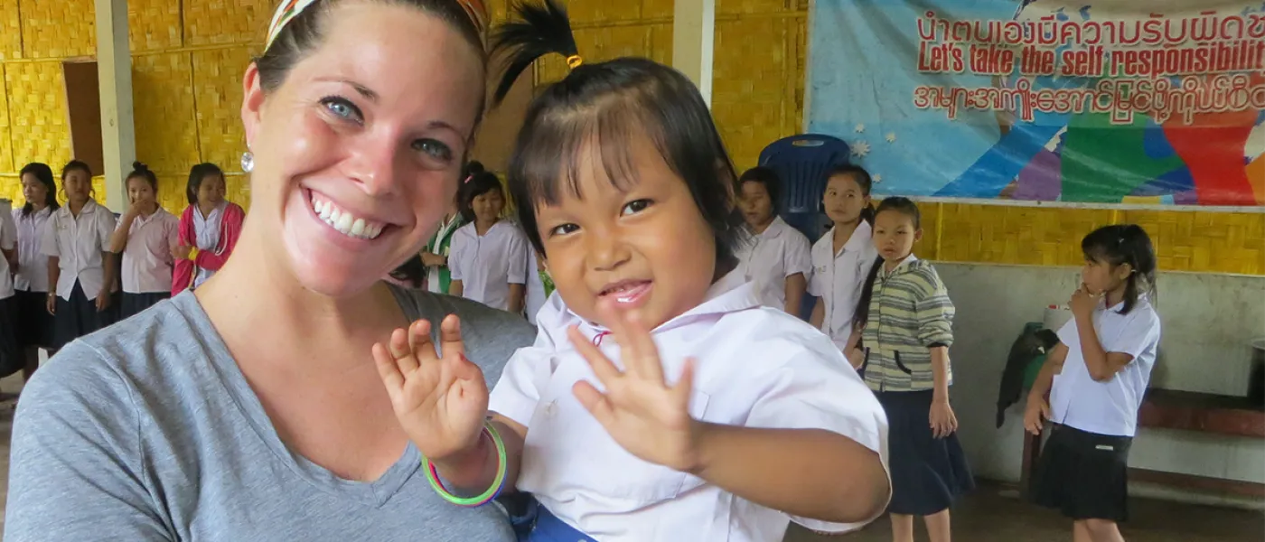 A student studying abroad in Thailand holds a baby who is waving for the camera