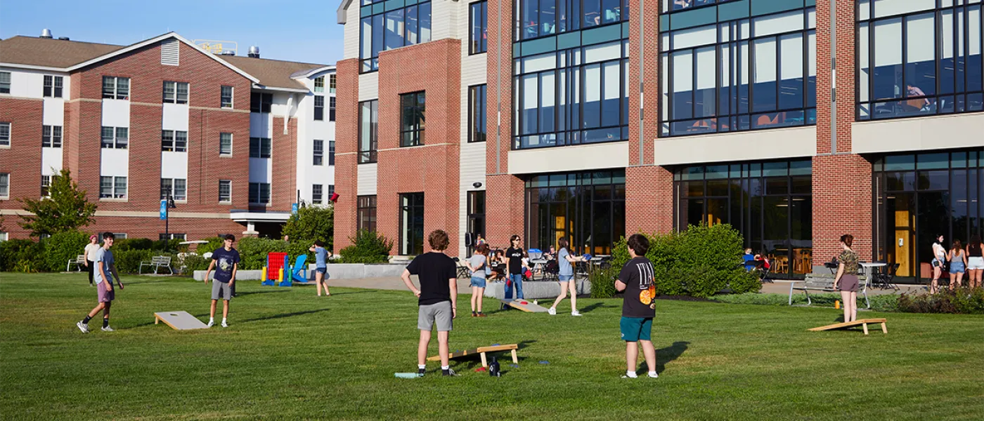 Several students play corn hole on green grass in front of a large brick building