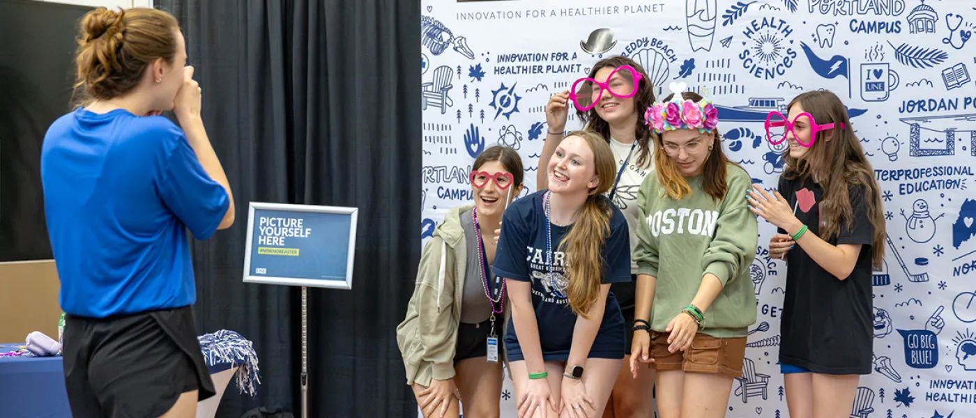 A student takes a photo of a group of students posing together in front of a wall made up of UNE-related graphics