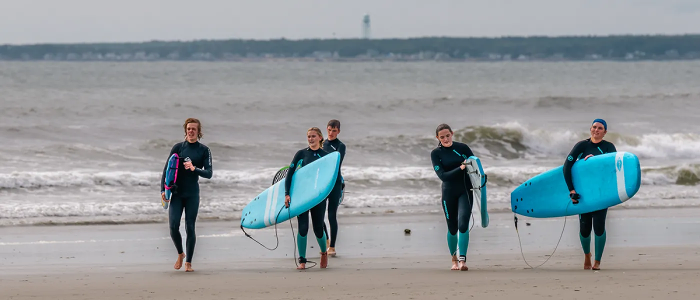 Five students walk on the beach away from the ocean holding their surfboards