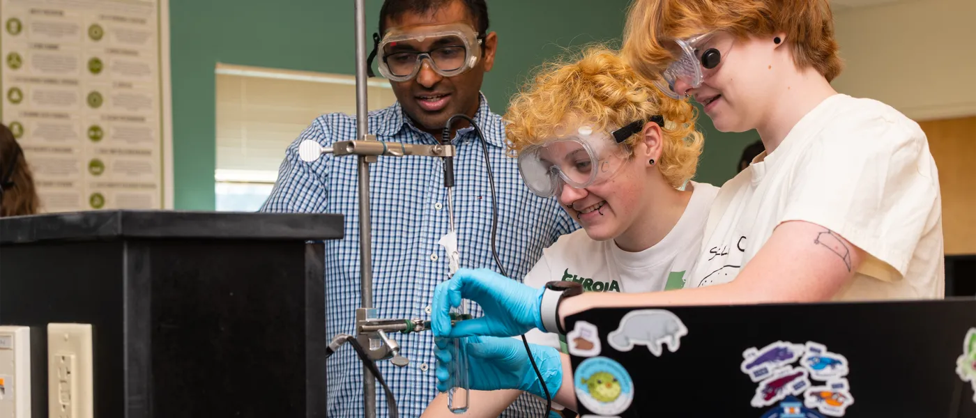Two students in safety glasses prep lab equipment as their professor guides them