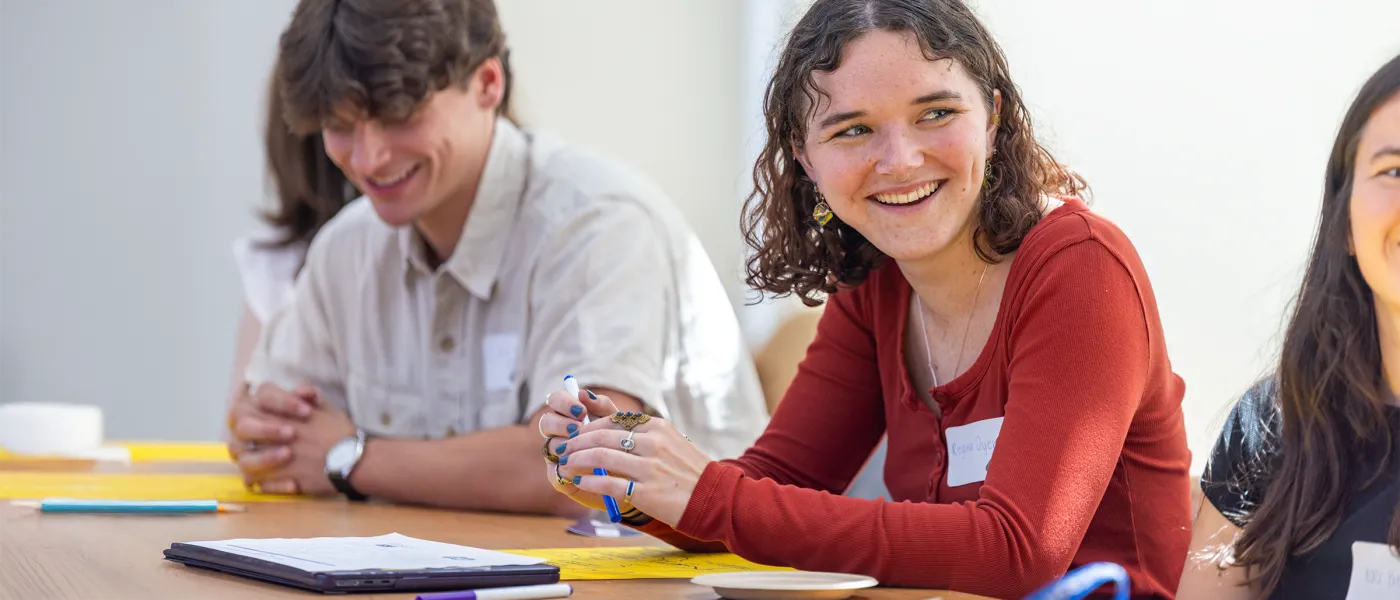A student smiles in a row of students sitting with papers and pencils laid on the long table in front of them