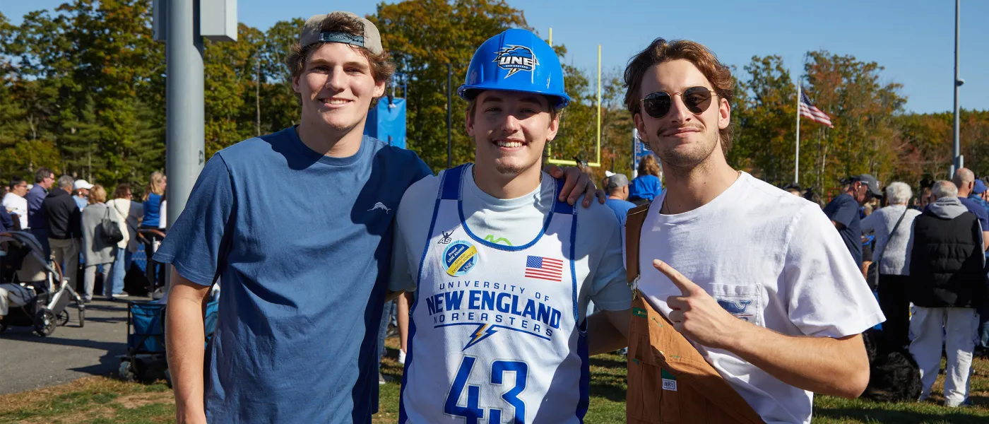 Three students pose for the camera, the middle student wearing a U N E hardhat and sports vest with "University of New England #43" and an alumni sticker
