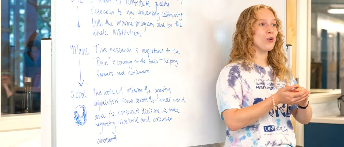 A student stands in front of a white board that has thoughts on UNE, Maine, and global education from the student written in blue marker