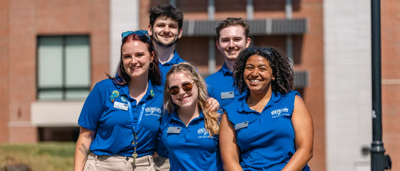 A group of orientation leaders stand together wearing matching blue polo shirts in front of a brick building