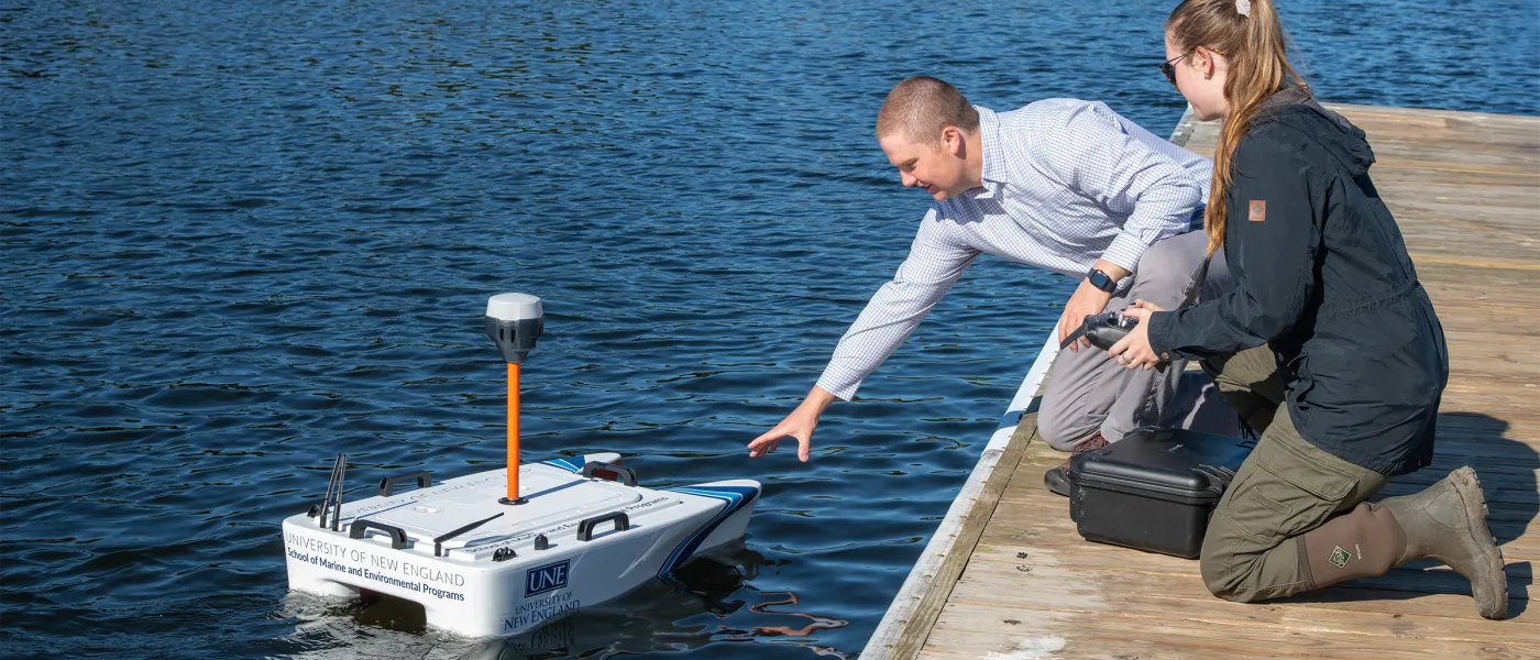 Two students send off a remote-controlled catamaran into a river from a wooden dock