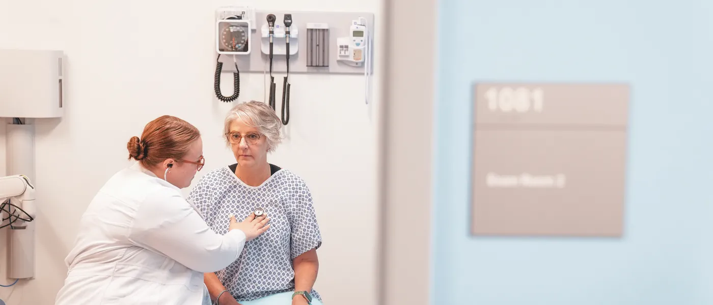 U N E C O M student using stethoscope to examine patient in hospital gown in clinical examination room.