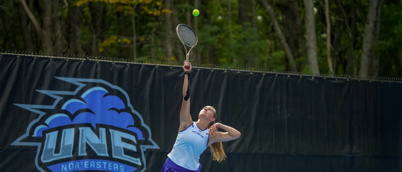 A U N E women's tennis player jumps to serve in front of the Nor'easters logo at the state-of-the-art outdoor tennis facility.
