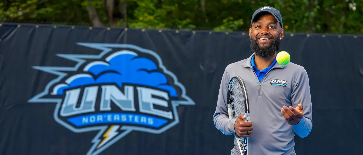 Coach Jovan Jordan-Whitter smiles while tossing a tennis ball in the air in front of the UNE Nor'easters logo at the Blue Cyclone Complex.