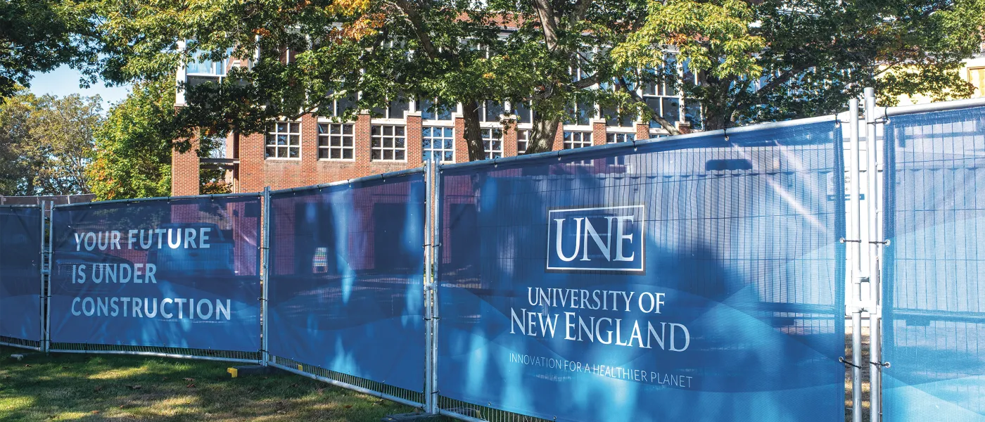 Blue construction fence displaying UNE branding and 'Your Future Is Under Construction' message in front of brick academic building with trees.