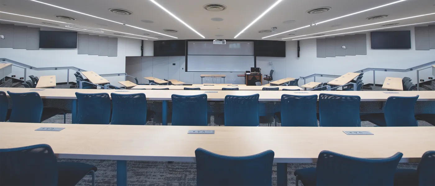Modern tiered lecture hall with blue chairs, wooden desks, projection screen, and linear ceiling lighting at UNE.