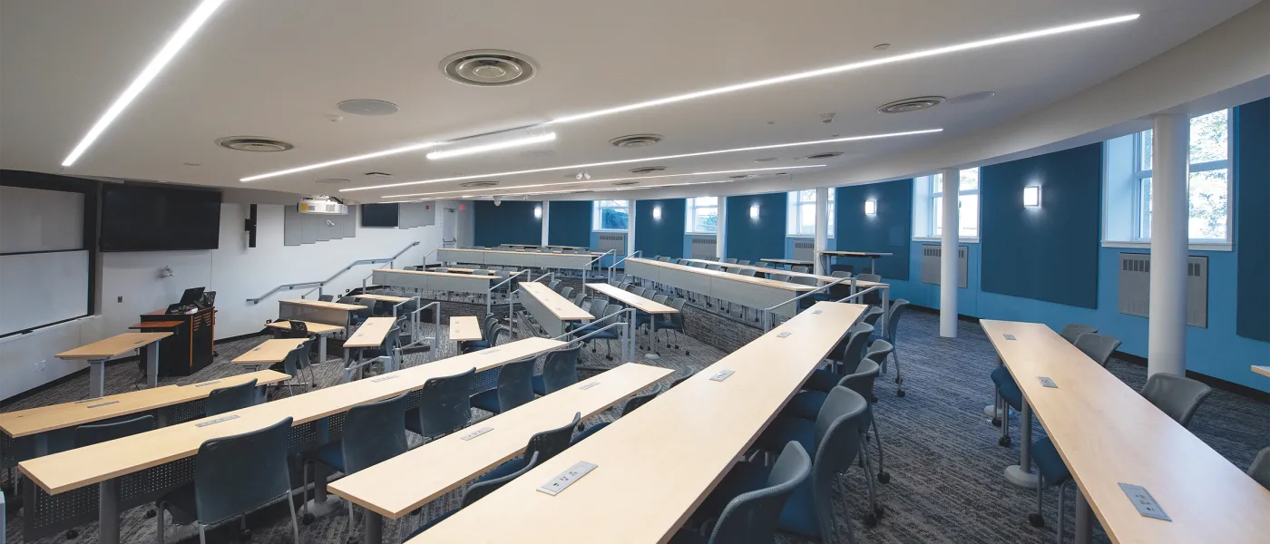 Contemporary classroom with rows of blue chairs, blonde wood desks, blue accent walls, and large windows providing natural light.