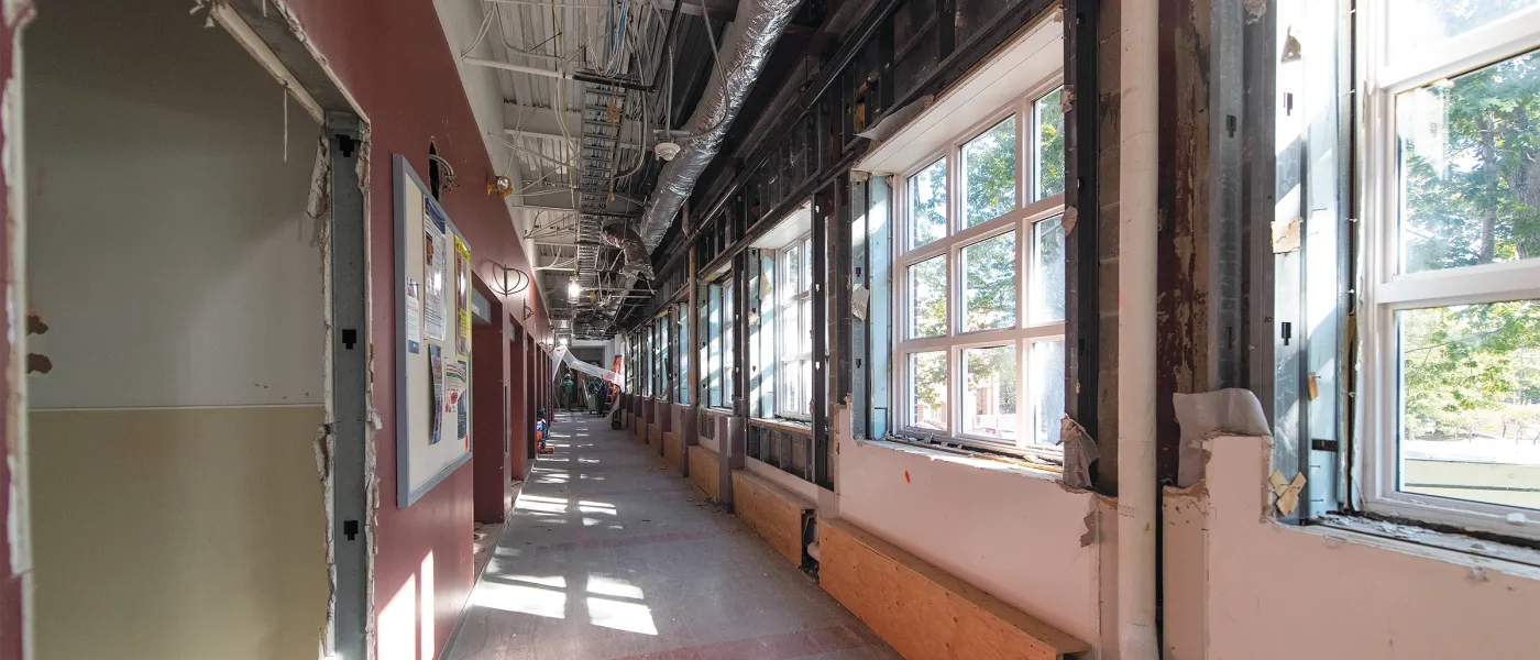 Long corridor under renovation with exposed ceiling ductwork, pink walls, construction materials, and windows along one side.