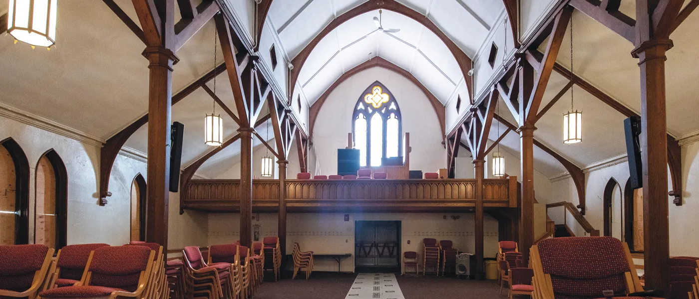 Interior of Ludcke featuring Gothic arched ceiling with dark wooden beams, arched windows, choir loft, and rows of red upholstered chairs.