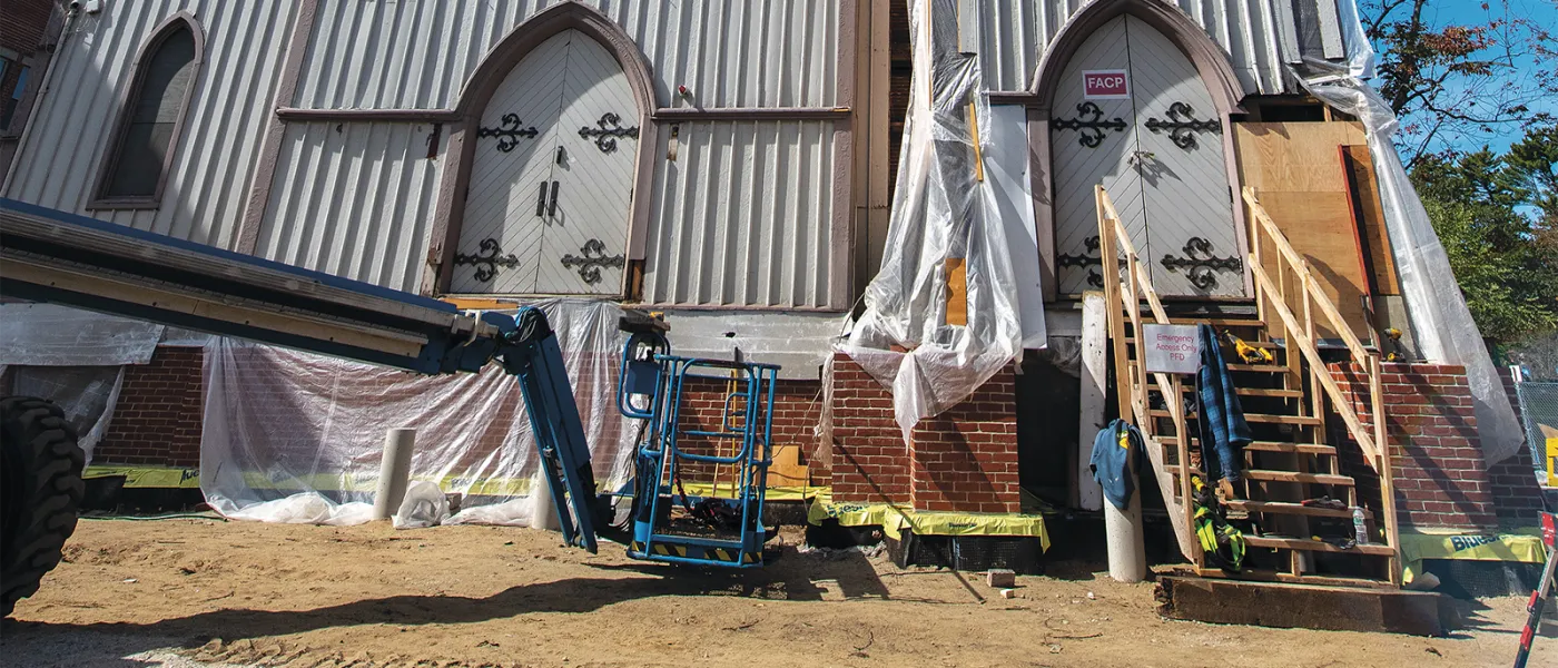 Construction workers on lift near Gothic arched entrance of white church building with protective sheeting and wooden scaffolding.