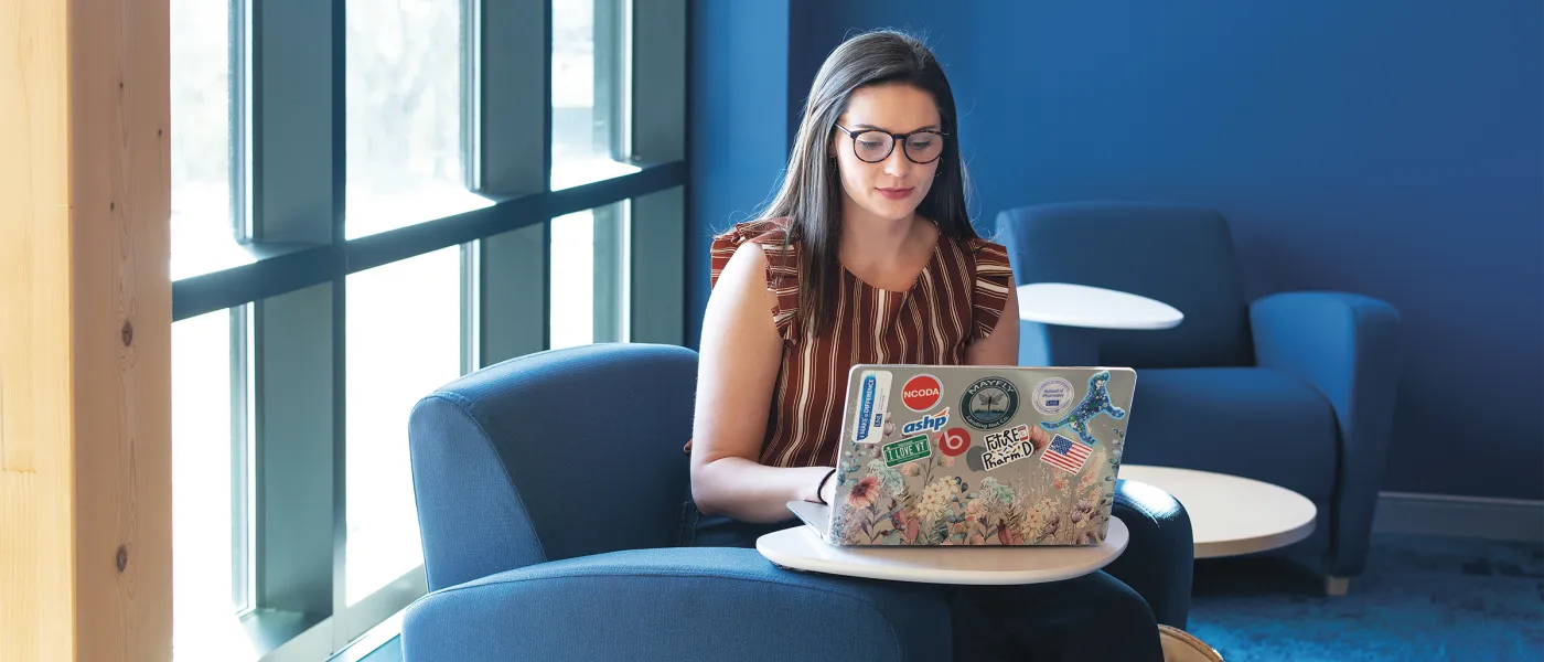 Student working on laptop covered with stickers while sitting in blue lounge chair by window.