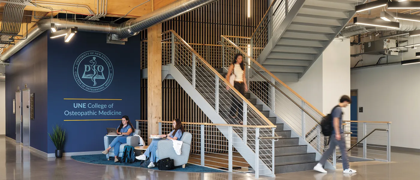 Students studying on white chairs near UNE College of Osteopathic Medicine branded wall beneath modern staircase.