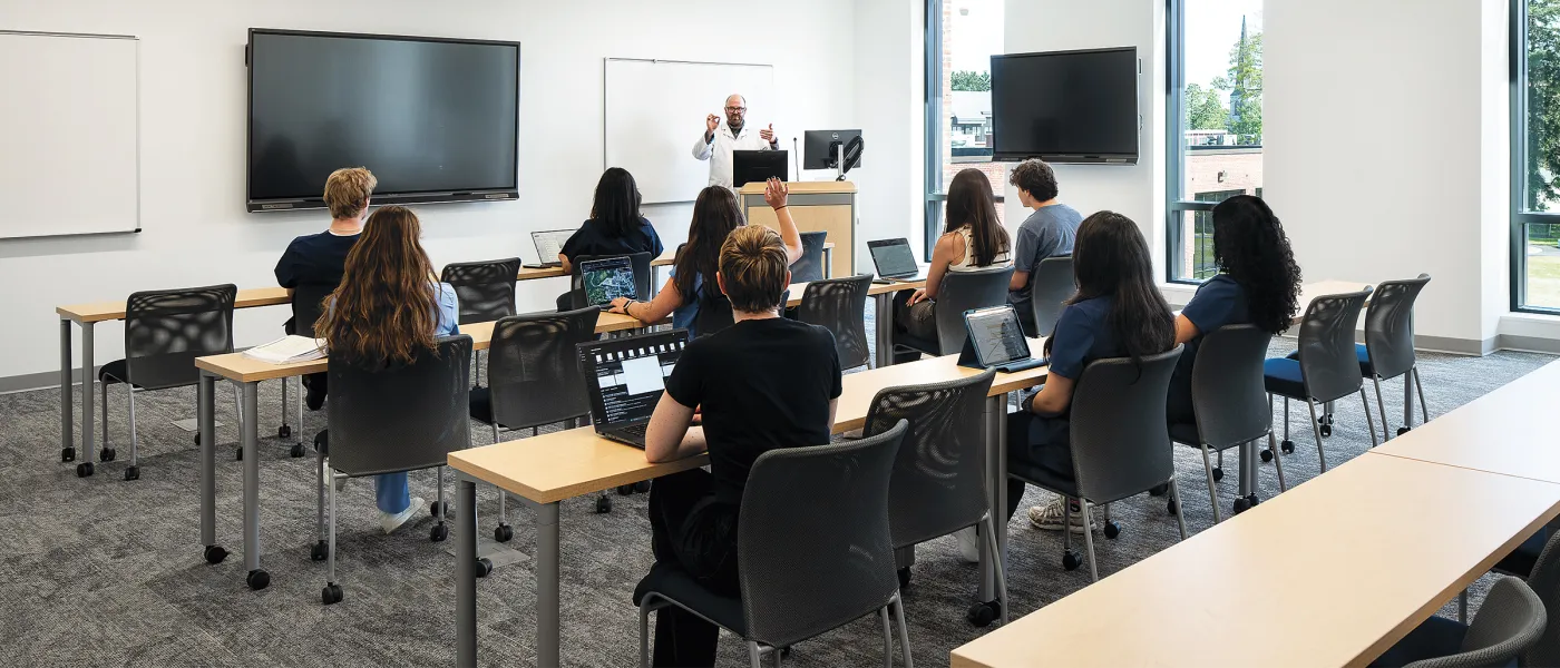 Instructor in white coat teaching students seated at tables with laptops in bright classroom with whiteboards and monitors.
