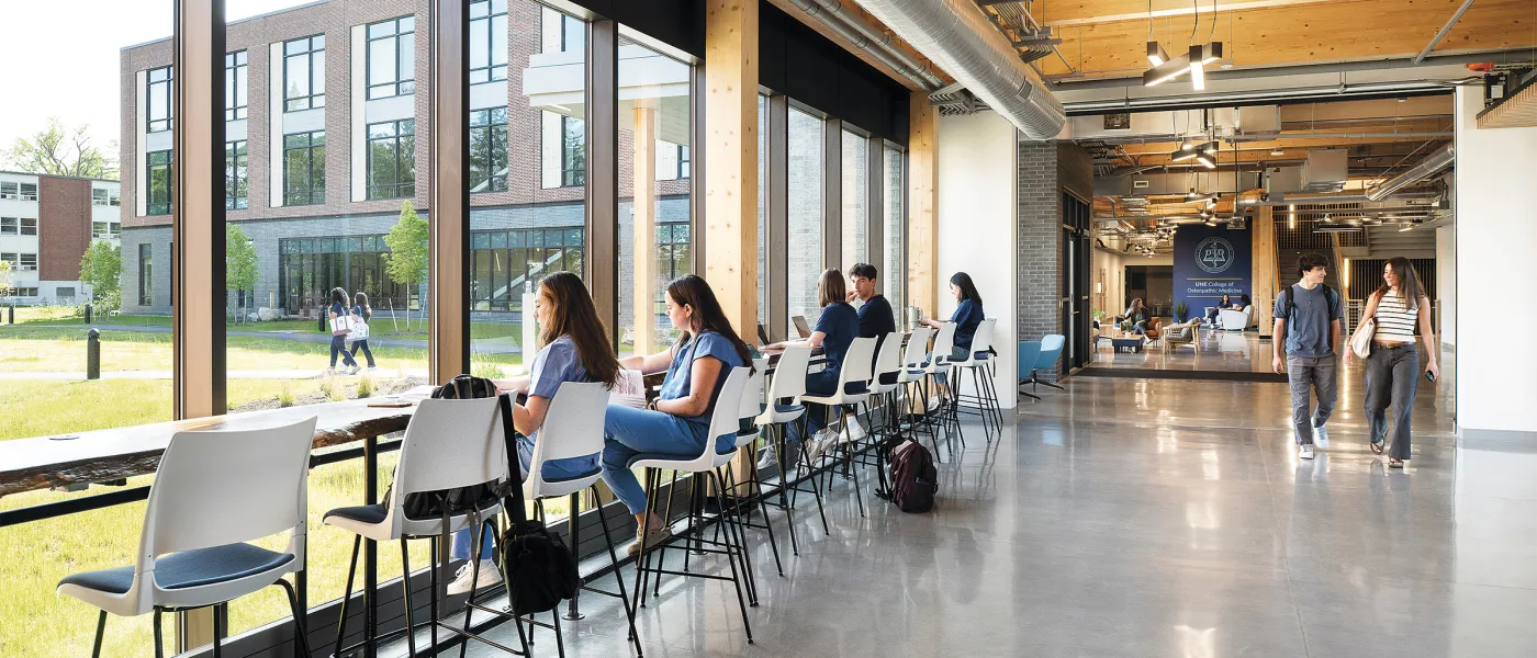 Students seated at counter-height chairs along window wall overlooking campus courtyard in bright hallway.