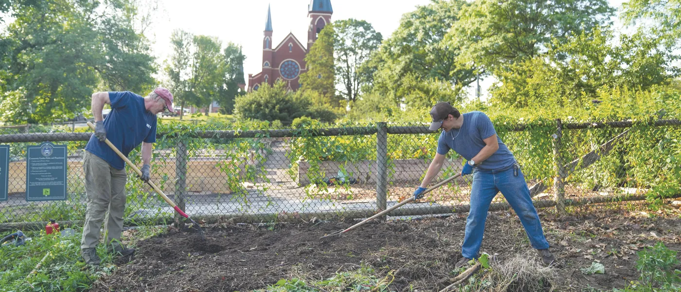 Luke Jenkins (Biology, ’26) and Cameron Wake, Ph.D. rake and prepare soil in a community garden for a composter with Cathedral of the Immaculate Conception visible in the background.
