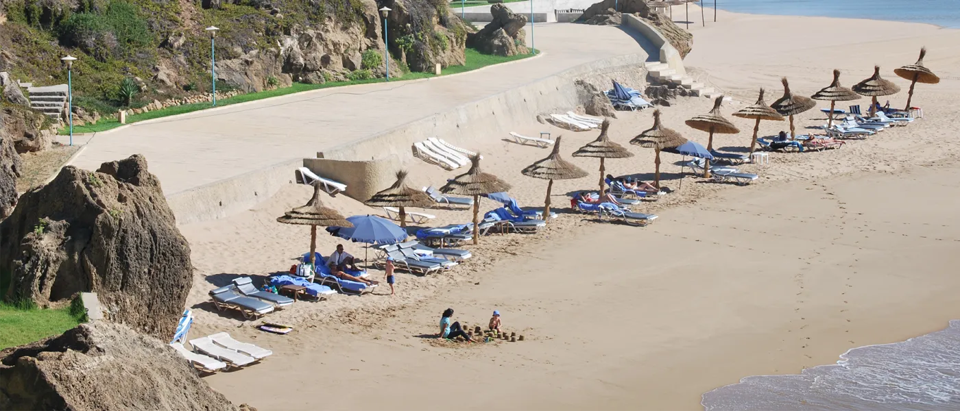 Sandy beach in Tangier with traditional thatched umbrellas, blue beach chairs, rocky outcroppings, and beachgoers along the Mediterranean coastline.
