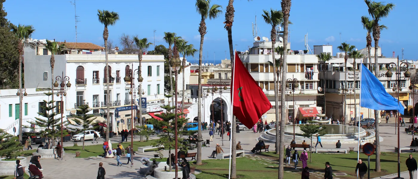 Public square in Tangier with Moroccan and French flags, palm trees, white colonial architecture, pedestrians, and traditional archway entrance to medina.