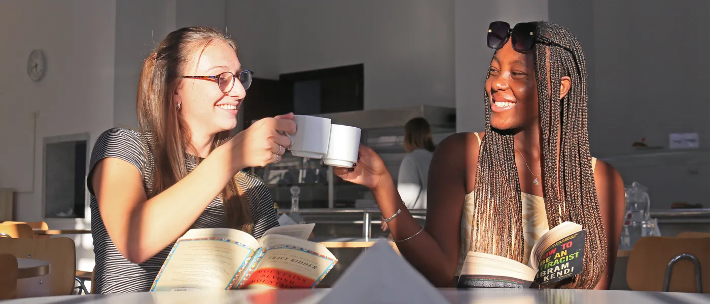 Two students toast coffee mugs while studying together at table with open books in bright modern common space.