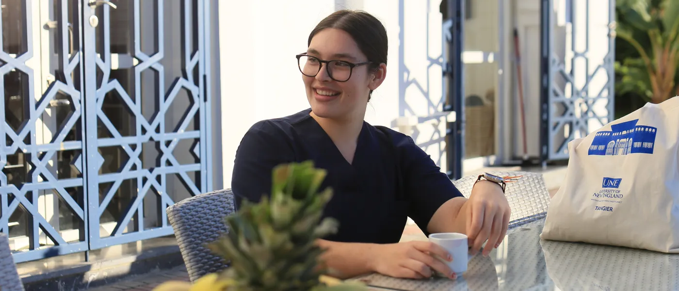 Student sits at outdoor café table with coffee and UNE Tangier tote bag, decorative blue metalwork visible in background.