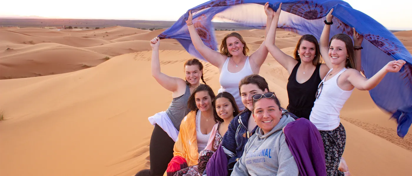 Group of students pose together on sand dune holding colorful scarves overhead during desert excursion at sunset.
