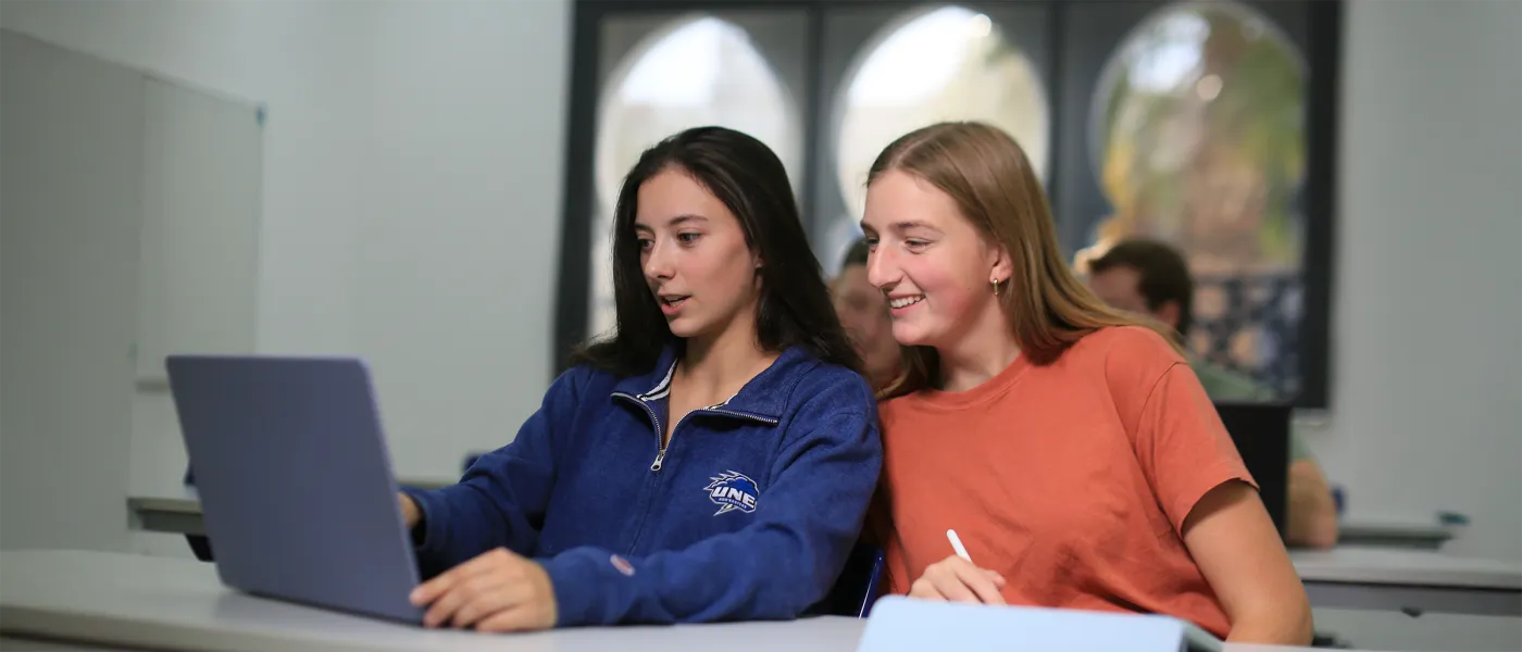 Two students study together at table with laptop and notebooks, Moroccan-style arched windows visible in background.